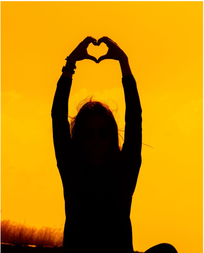 A silhouette raising her arms in a heart shape against a golden sunset sky
